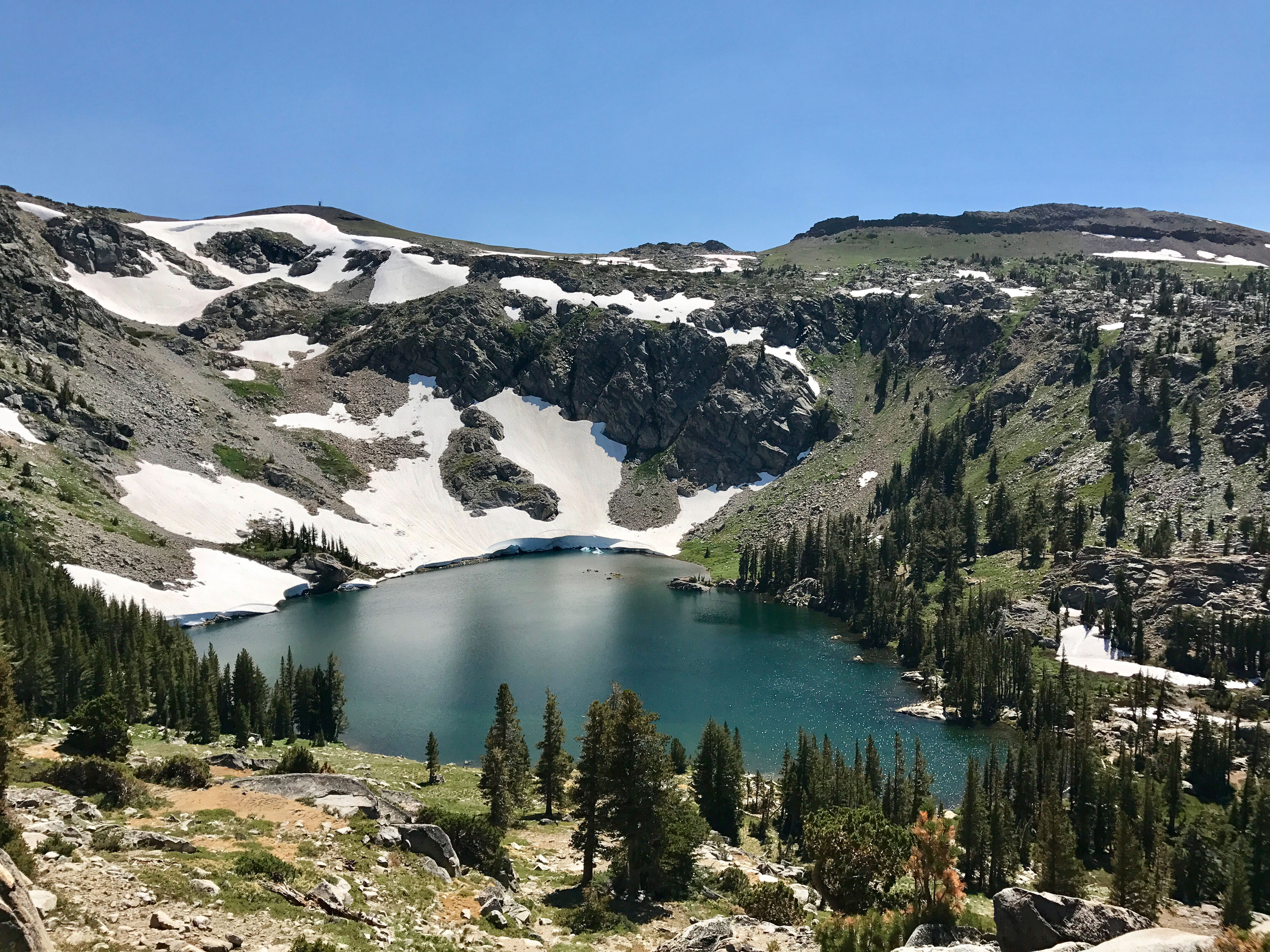View from above of an aqua Blue Lake surrounded by lush green trees and rocks and snow